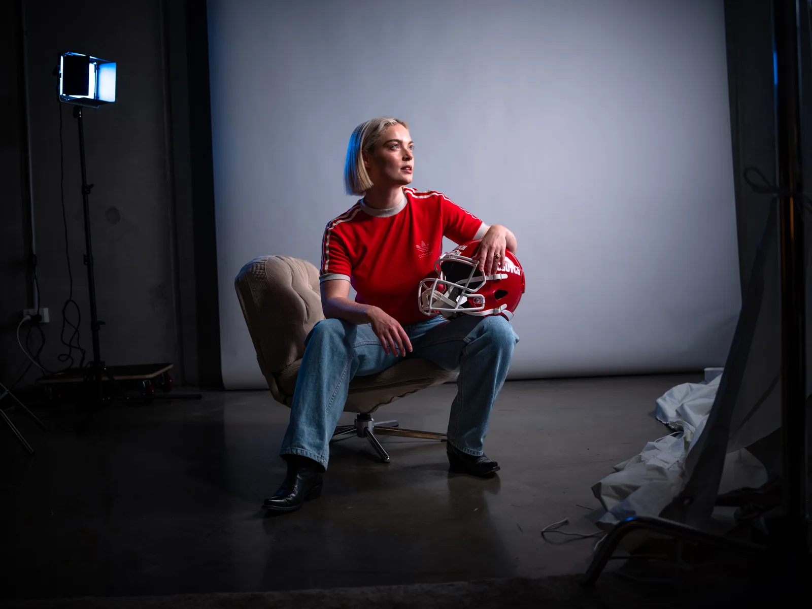 Lucy in a red tee and jeans, sitting on a studio chair with a red CLUTCH football helmet under her arm