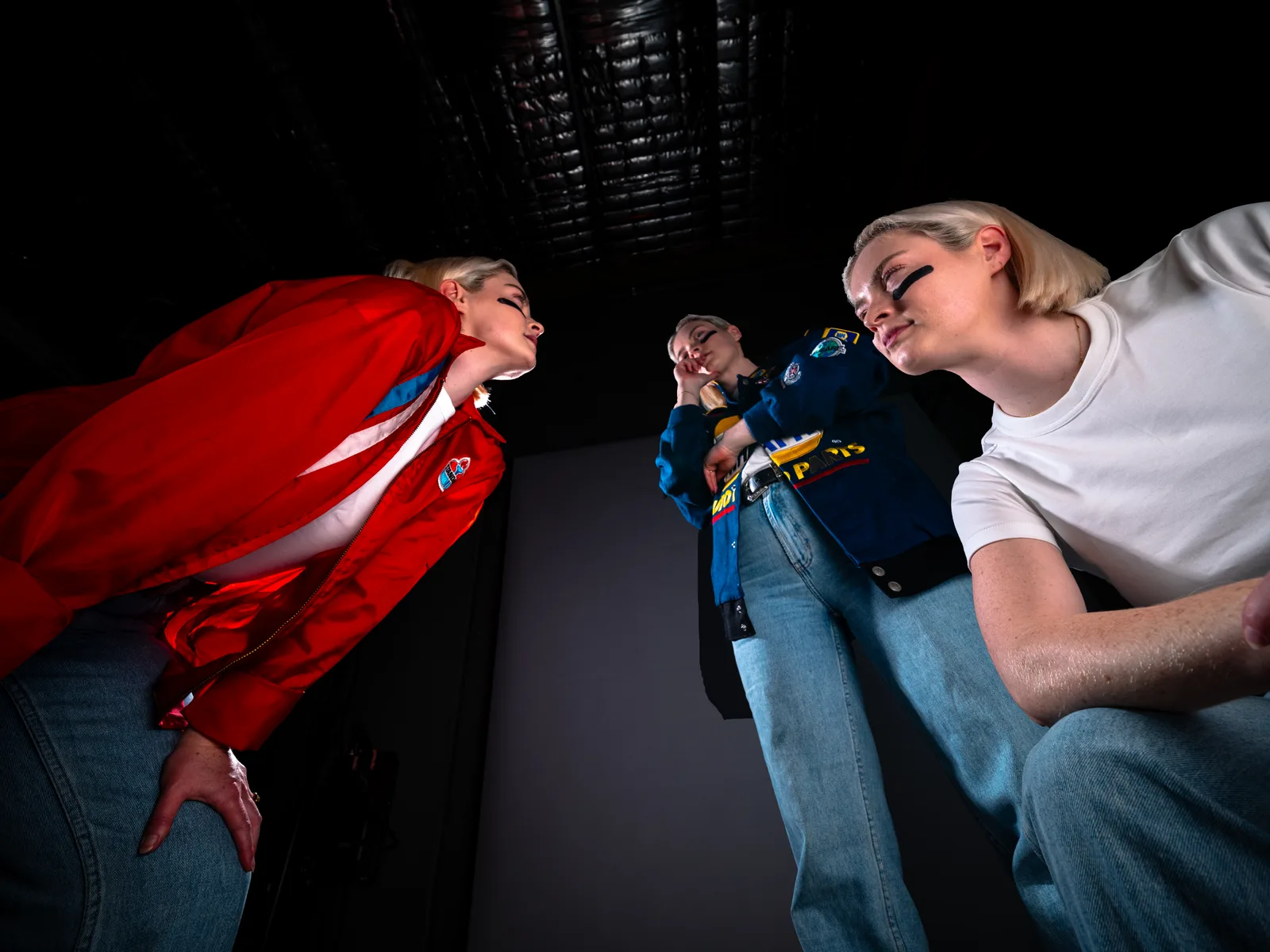 Three Lucys from a low angle in the studio: one in a red jacket, one in a NAPA racing jacket, one in a white tee with eye black