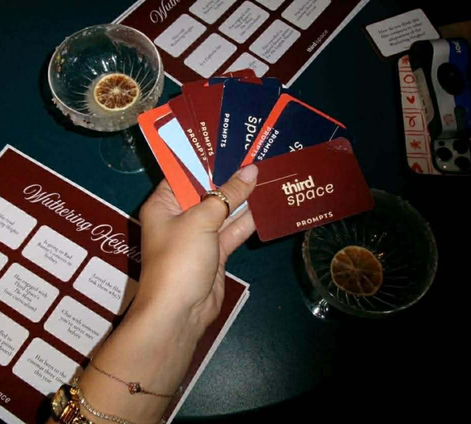 A hand fanning out Third Space prompt cards over a Wuthering Heights bingo sheet and two cocktails